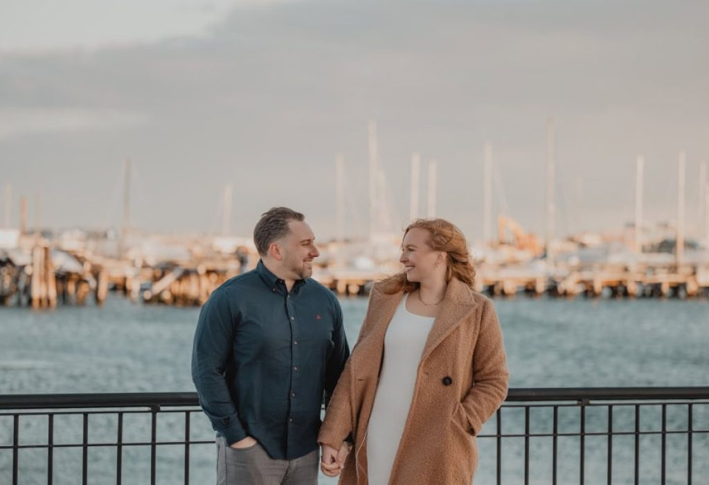 Couple standing by Boston Harbor holding hands and smiling at each other
