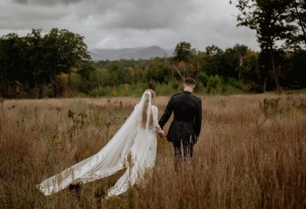 Bride with flowing veil and groom walking hand in hand through tall grass with mountain view in NH