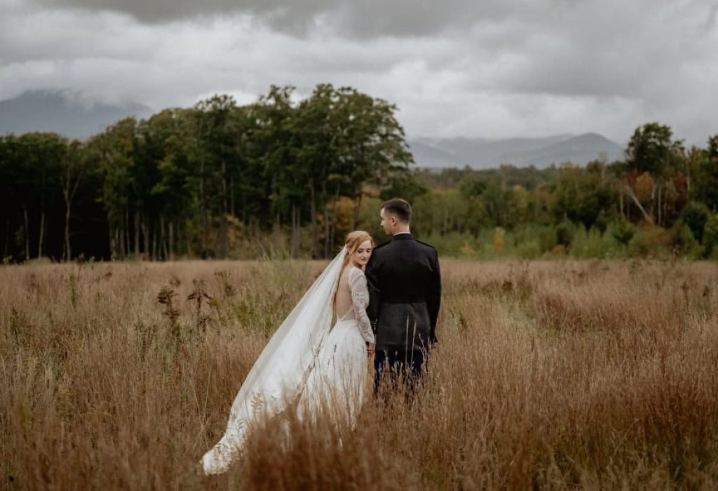Bride looking over shoulder at groom while standing in tall grass at scenic outdoor New Hampshire wedding