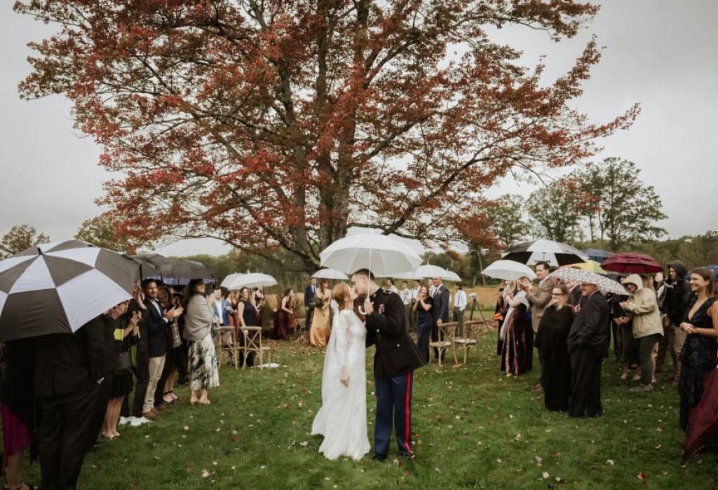 Bride and groom sharing first kiss under umbrella as guests cheer during fall wedding ceremony in New Hampshire