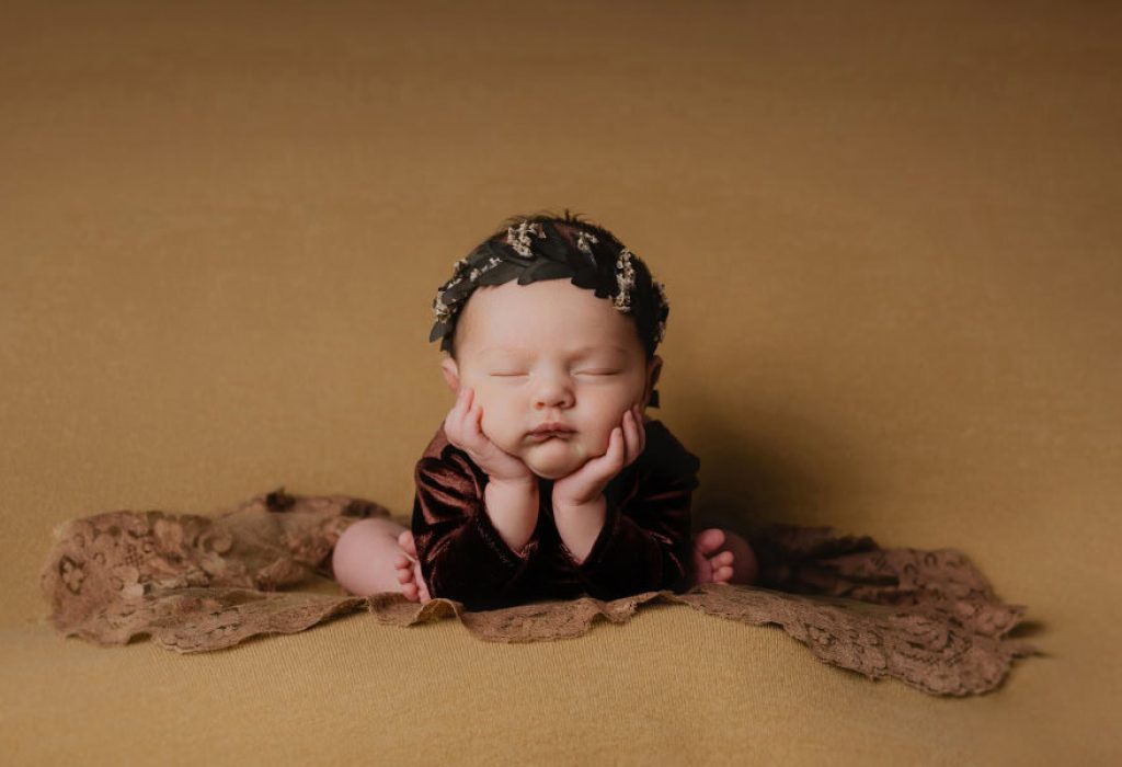 Newborn baby girl posed in classic froggy position on mustard backdrop wearing brown velvet outfit and headband