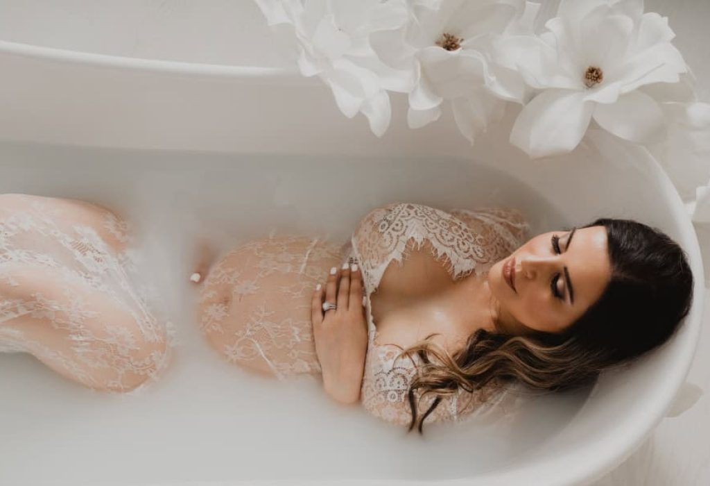 Pregnant woman in lace robe standing beside a white clawfoot tub with soft floral decor during studio maternity session in New Hampshire