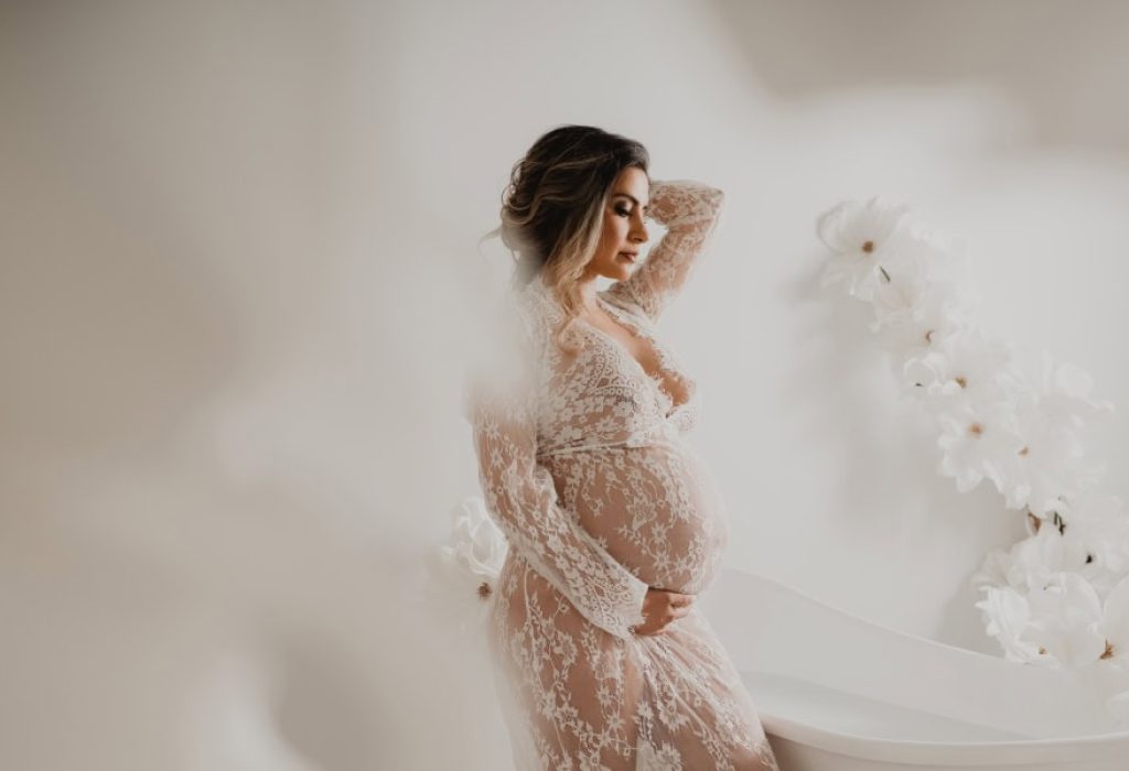 Pregnant woman in lace robe standing beside a white clawfoot tub with soft floral decor during studio maternity session in New Hampshire