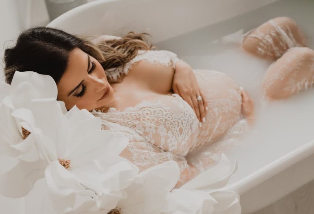 Mother-to-be relaxing in a milk bath wearing a sheer lace gown, surrounded by large white florals in a light-filled maternity photo studio