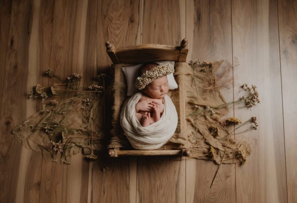 Wrapped newborn baby posed in rustic wooden bed with floral crown and dried flowers on wood floor