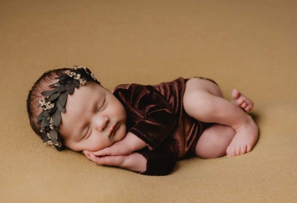 Newborn posed on side, asleep on mustard blanket in soft brown velvet outfit and dark floral headband