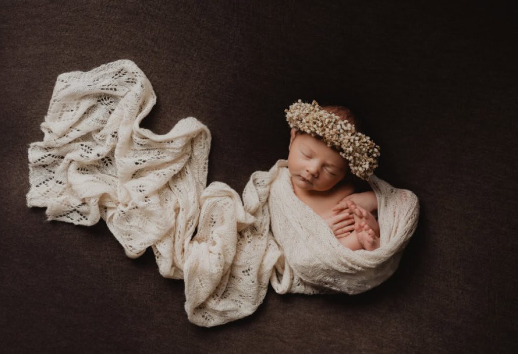 Newborn baby swaddled in ivory wrap with knit bonnet and delicate floral crown on brown background