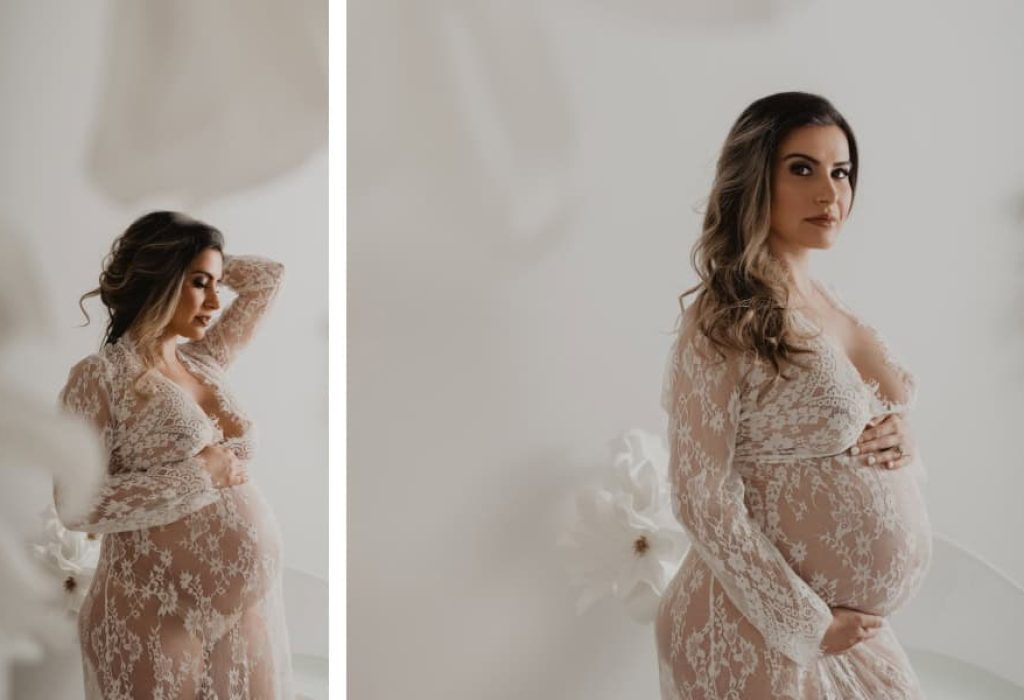 Studio maternity portrait of woman in lace robe holding her belly with white florals and soft, minimalist background